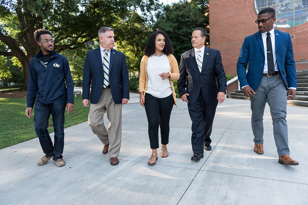 Five people walking across campus in a group. Three of them are black students. The person in the center is a woman, and the other four look at her while she speaks.