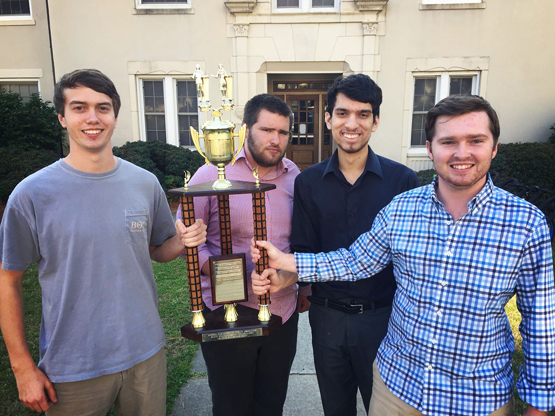 Four young men outside a campus building with a trophy