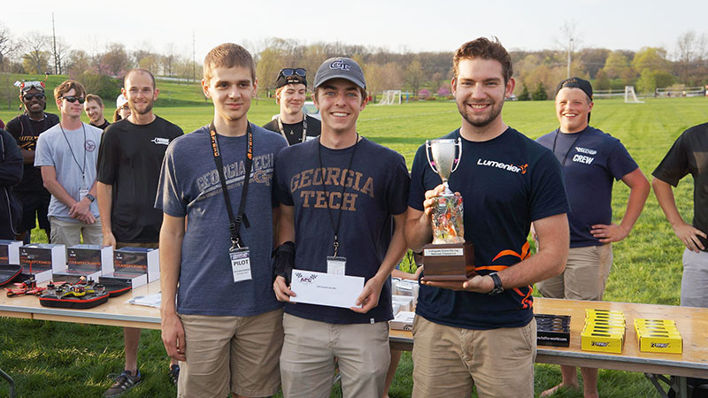 The three men of the Tech drone racing team holding their awards and smiling with drone racing crew and other teams behind them