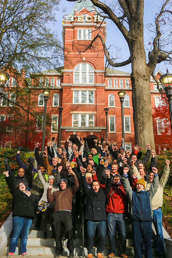 About two dozen people of various ages standing in a large group at the base of Tech Tower are happily lifting their arms in victory and smiling.