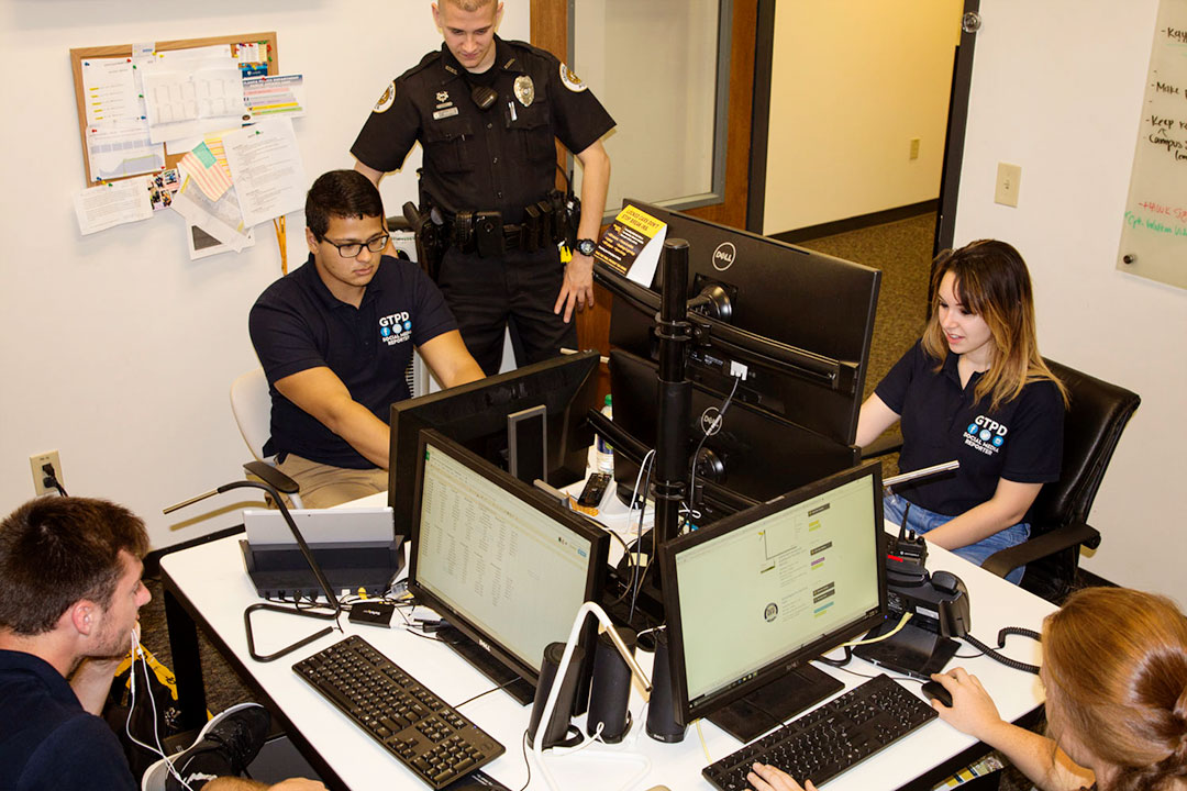 A police officer watches while four members of the GTPD social media team work at a group of computers