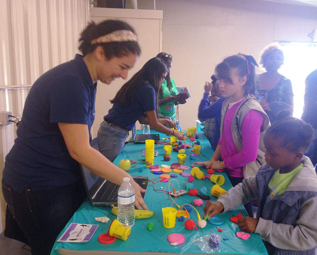 A teenage woman teaching some girls under age 10 with modeling clay, wires and a computer