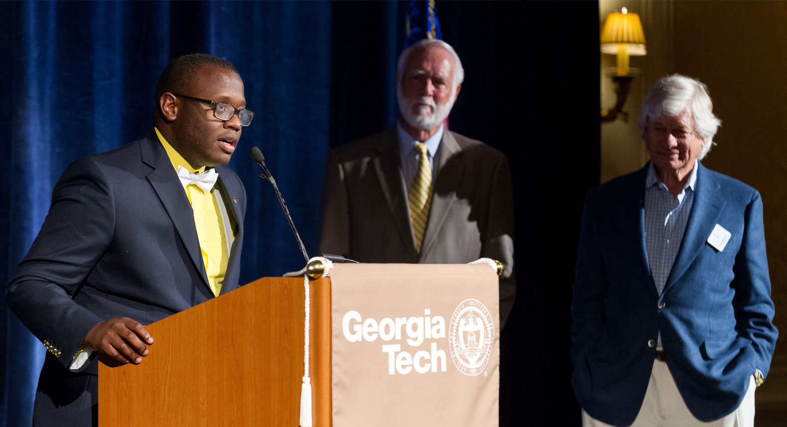 Duane Carver speaking on stage with G. Wayne Clough and Al West standing nearby