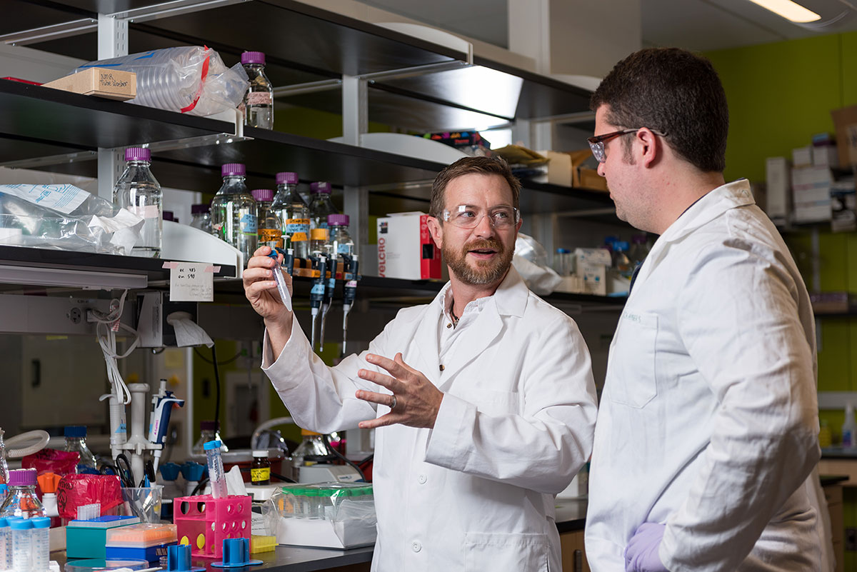 Two men in laboratory, wearing lab coats and safety glasses, with one holding up a test tube for the other to see