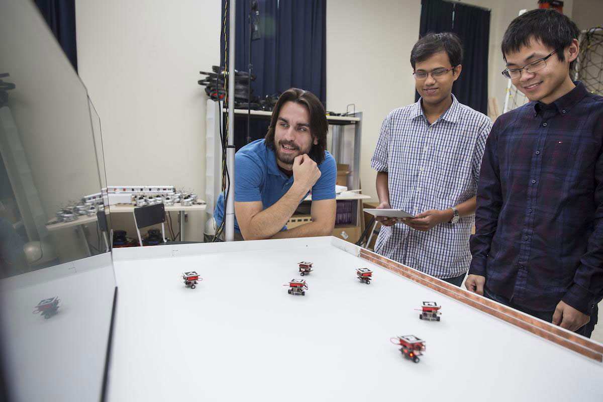 Three students working with small, palm-sized robots on a table in the Georgia Tech Robotarium