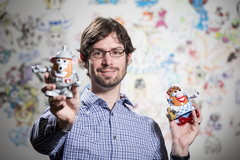 Mark Riedl holding up Mr. and Mrs. Potato Head figures in front of a colorful, child-like designed wall