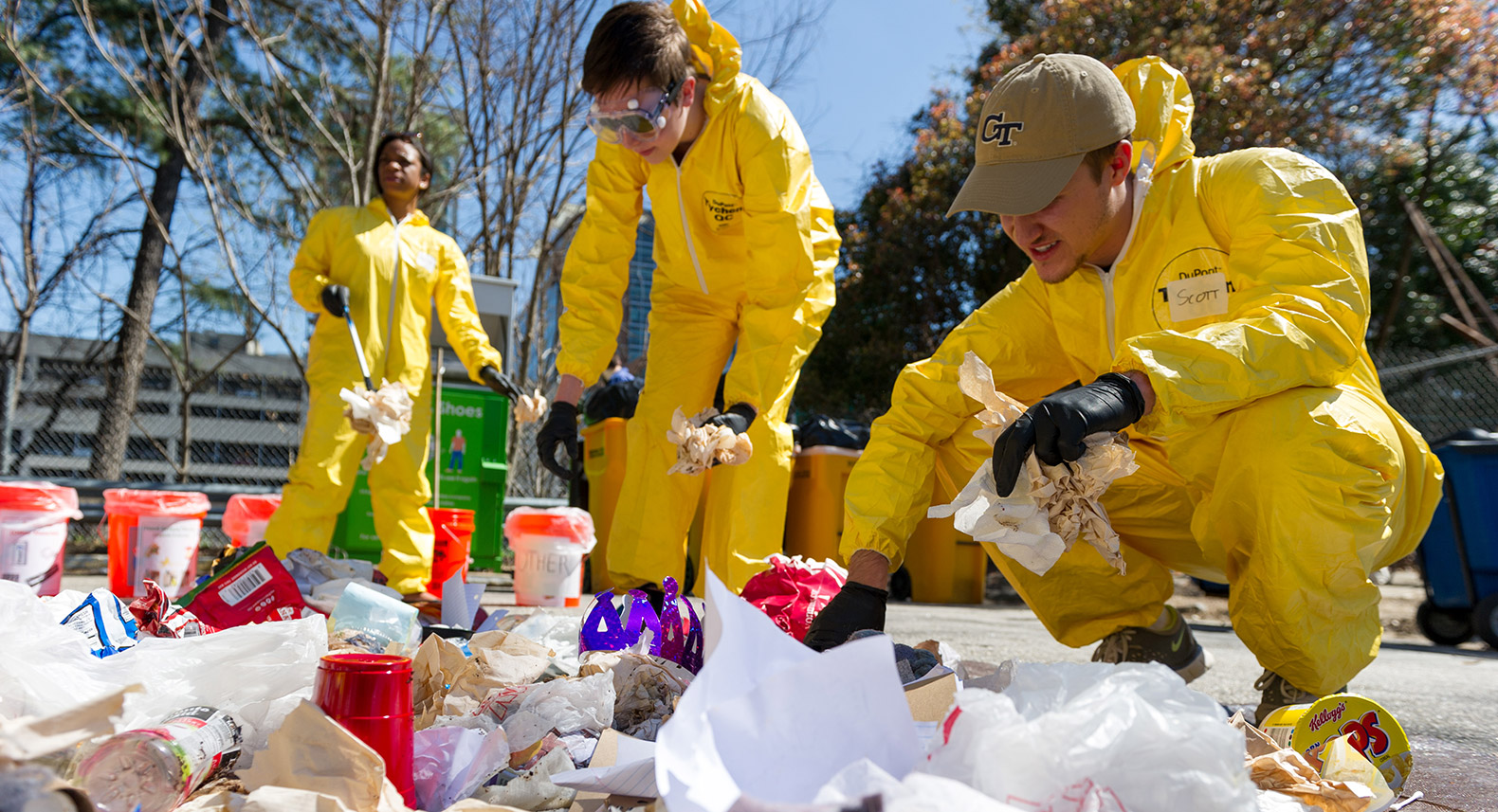 Students in yellow plastic jumpsuits dig through a pile of trash on the ground and sort it into orange 5-gallon buckets labeled with categories of recycling and waste