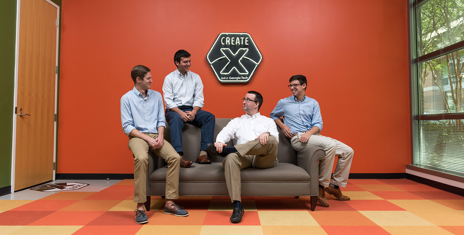 Portrait of the four TEQ charging members sitting on a couch under a black-and-white Create-X neon sign on an orange wall